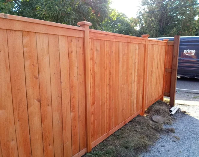 A newly installed light brown wooden fence stands beside a gravel and grassy area, with an Amazon Prime truck in the background.